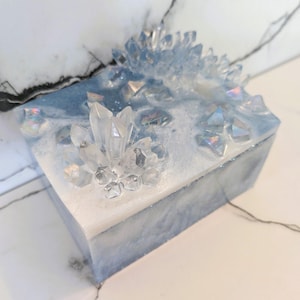 May include: A square, decorative box with a white and blue marbled design. The top is adorned with clear, crystal-like formations of varying sizes. The box has a glittery, iridescent finish, giving it a magical appearance.