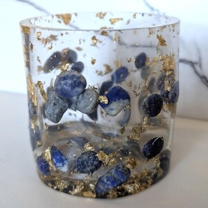 May include: A clear, cylindrical glass with embedded blue stones and gold leaf flakes. The stones vary in size and shade, creating a textured appearance. The gold leaf adds a touch of luxury to this decorative item.