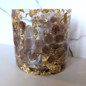 Puede incluir: Un recipiente cilíndrico transparente de resina con pan de oro incrustado y pequeñas piedras redondas y ovaladas en tonos marrones y blancos. El pan de oro se concentra en la parte inferior y se dispersa por la resina.