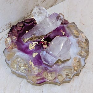 May include: A decorative resin coaster with a purple and white design, accented with gold leaf and clear quartz crystals. The coaster has a gold-colored edge and features several crystal formations.