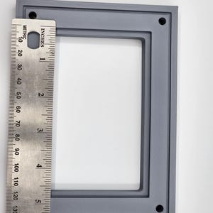 K&ouml;nnte beinhalten: Ein grauer rechteckiger Rahmen mit abgerundeten Ecken und vier L&ouml;chern, gezeigt mit einem Lineal. Der Rahmen ist etwa 15 cm hoch. Der Rahmen besteht aus einem festen Material und hat eine strukturierte Oberfl&auml;che.