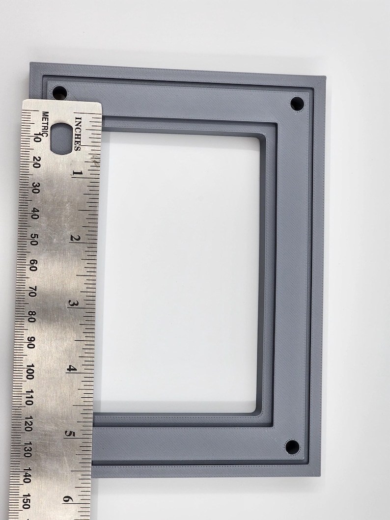 K&ouml;nnte beinhalten: Ein grauer rechteckiger Rahmen mit abgerundeten Ecken und vier L&ouml;chern, gezeigt mit einem Lineal. Der Rahmen ist etwa 15 cm hoch. Der Rahmen besteht aus einem festen Material und hat eine strukturierte Oberfl&auml;che.