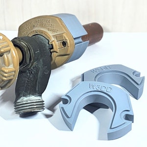 Könnte beinhalten: Nahaufnahme eines braunen und grauen Außenhahns mit Frostschutzventil. Der Hahn hat ein Gewindeende zum Anschließen eines Schlauches. Zwei graue Kunststoff-Schlauchschellen mit dem Text "W3DD" sind ebenfalls zu sehen.