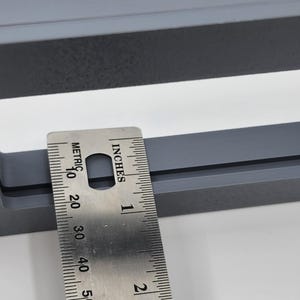 K&ouml;nnte beinhalten: Nahaufnahme eines grauen, rechteckigen Objekts mit einem Metalllineal, das Zoll- und metrische Ma&szlig;e anzeigt. Das Lineal liegt auf dem Objekt und zeigt dessen Abmessungen an. Das Objekt scheint ein Rahmen oder eine &auml;hnliche Struktur zu sein.