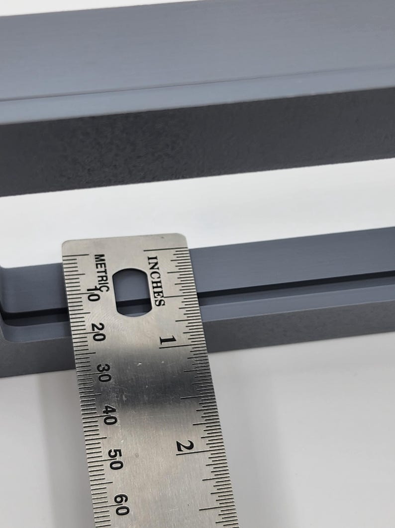 K&ouml;nnte beinhalten: Nahaufnahme eines grauen, rechteckigen Objekts mit einem Metalllineal, das Zoll- und metrische Ma&szlig;e anzeigt. Das Lineal liegt auf dem Objekt und zeigt dessen Abmessungen an. Das Objekt scheint ein Rahmen oder eine &auml;hnliche Struktur zu sein.