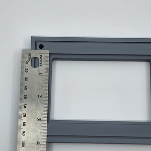 K&ouml;nnte beinhalten: Ein grauer rechteckiger Rahmen mit wei&szlig;em Innenraum, etwa 12,7 cm lang, wird neben einem Lineal gezeigt. Das Lineal zeigt sowohl Zoll als auch Zentimeter an, wobei die L&auml;nge des Rahmens markiert ist. Der Rahmen hat ein kleines Loch in der oberen linken Ecke.