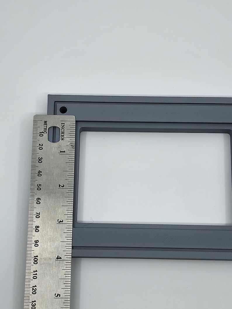 K&ouml;nnte beinhalten: Ein grauer rechteckiger Rahmen mit wei&szlig;em Innenraum, etwa 12,7 cm lang, wird neben einem Lineal gezeigt. Das Lineal zeigt sowohl Zoll als auch Zentimeter an, wobei die L&auml;nge des Rahmens markiert ist. Der Rahmen hat ein kleines Loch in der oberen linken Ecke.