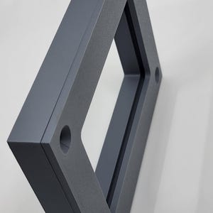 K&ouml;nnte beinhalten: Ein dunkelgrauer, geometrischer Rahmen mit einer rechteckigen &Ouml;ffnung. Der Rahmen hat eine strukturierte Oberfl&auml;che und zwei kreisf&ouml;rmige L&ouml;cher. Das Objekt ist vor einem wei&szlig;en Hintergrund abgewinkelt, wodurch Schatten entstehen.