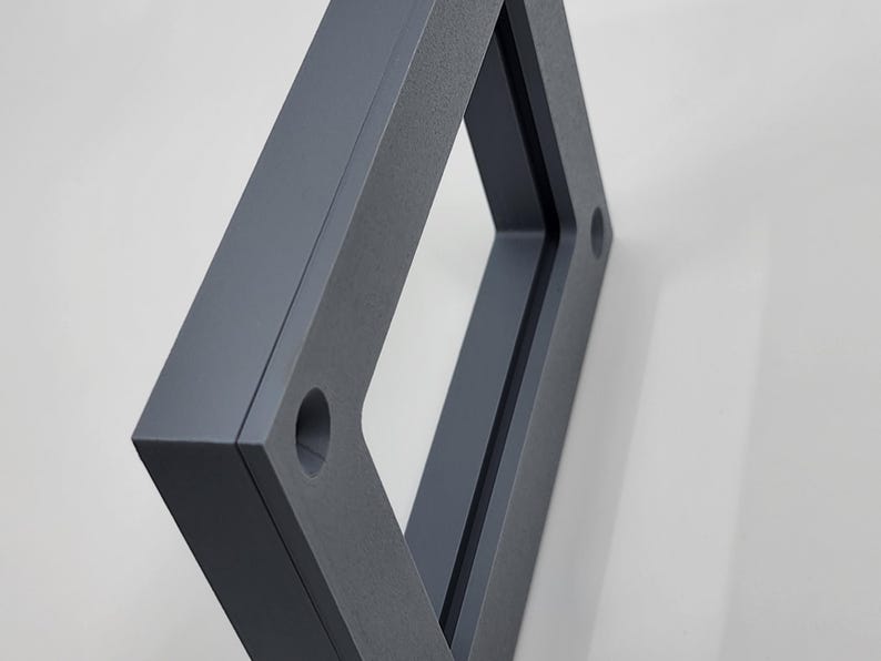 K&ouml;nnte beinhalten: Ein dunkelgrauer, geometrischer Rahmen mit einer rechteckigen &Ouml;ffnung. Der Rahmen hat eine strukturierte Oberfl&auml;che und zwei kreisf&ouml;rmige L&ouml;cher. Das Objekt ist vor einem wei&szlig;en Hintergrund abgewinkelt, wodurch Schatten entstehen.