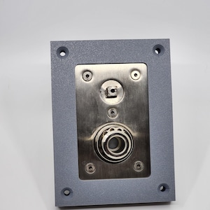 K&ouml;nnte beinhalten: Eine graue rechteckige Metallplatte mit einem Mittelteil aus Edelstahl. Die Mitte weist einen kreisf&ouml;rmigen Mechanismus mit einer Reihe konzentrischer Ringe auf. Vier Schrauben befestigen die Edelstahlplatte am grauen Rahmen. Vier L&ouml;cher befinden sich in den Ecken.