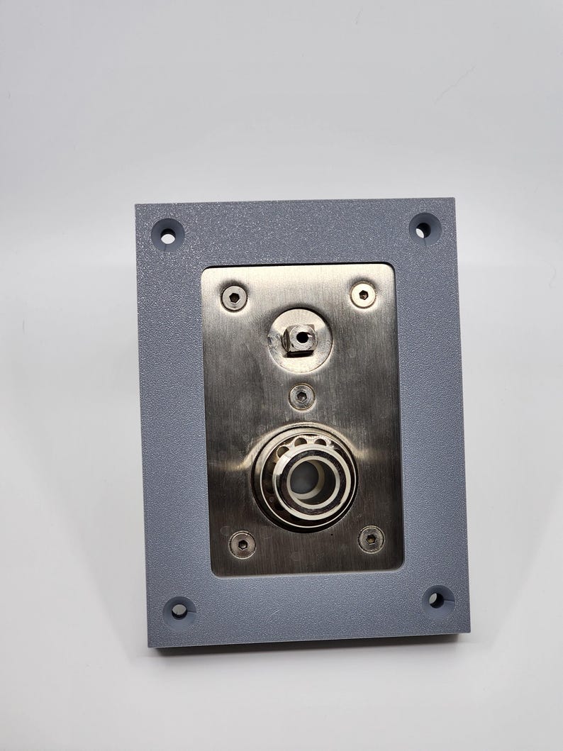 K&ouml;nnte beinhalten: Eine graue rechteckige Metallplatte mit einem Mittelteil aus Edelstahl. Die Mitte weist einen kreisf&ouml;rmigen Mechanismus mit einer Reihe konzentrischer Ringe auf. Vier Schrauben befestigen die Edelstahlplatte am grauen Rahmen. Vier L&ouml;cher befinden sich in den Ecken.