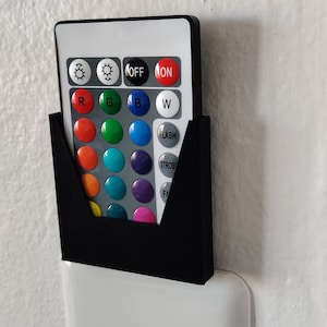 Puede incluir: Un interruptor de luz blanco con un soporte negro para mando a distancia montado encima. El mando a distancia tiene una variedad de botones de colores y el texto "ON", "OFF", "FLASH", "STROBE" y "FADE".