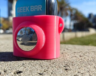 Op de afbeelding: Een felroze beschermende siliconen hoes met daarin een blauw en zilverkleurig Geek Bar Pulse wegwerp-vape-apparaat. De hoes heeft een ronde uitsparing aan de voorzijde, waarin een reflecterende zilveren cirkelvormige inzet zichtbaar is.