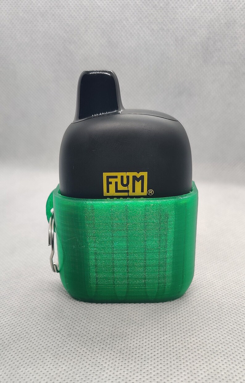 May include: A black and green electronic device with the word "FLUM" printed on the front. The device has a small, black mouthpiece and a green, textured case with a metal ring.