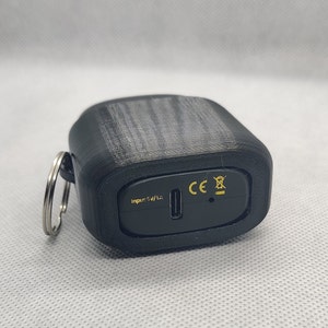 May include: Black charging case with a USB-C port and a keychain loop. The case has the text "Input: 5V/1A" and the CE mark.