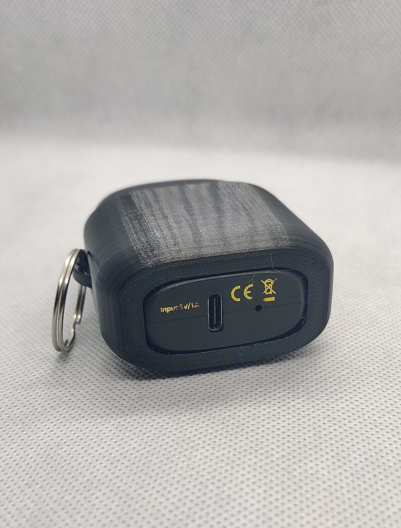 May include: Black charging case with a USB-C port and a keychain loop. The case has the text "Input: 5V/1A" and the CE mark.