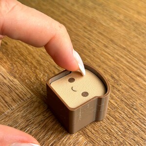 Op de afbeelding: Een klein, bruin, kubusvormig object met een lichtbruin, broodvormig inzetstuk aan de bovenkant. Het beige oppervlak is voorzien van een donkerbruine print van twee ronde ogen en een gebogen lijn als mond, lijkend op een stukje toast.