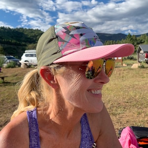 May include: A woman wearing a pink and green patterned baseball cap with a pink brim and reflective sunglasses. She is smiling and looking to the right.