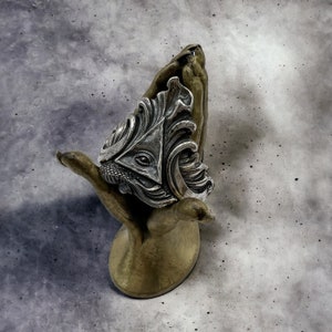 May include: A silver ring with an intricate design featuring a triangle with an eye inside, surrounded by swirling patterns. The ring is displayed on a gold hand-shaped stand.