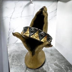 May include: A gold and black bracelet with a repeating pattern of stylized faces. The bracelet is displayed on a gold hand sculpture.