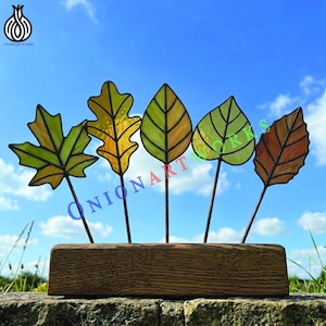 Puede incluir: Cinco decoraciones de hojas de vidriera en tonos verdes, amarillos y naranjas están montadas sobre finas varillas metálicas y colocadas en una base de madera. El fondo es un cielo azul. El texto "ONIONARTWORKS" es visible.