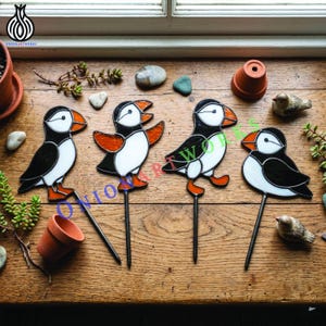 Puede incluir: Cuatro estacas de jardín de vitral con forma de frailecillos, hechas con piezas de vidrio negro, blanco y naranja con líneas de soldadura de metal. Cada ave está unida a una varilla de metal. El texto ONION ARTWORKS se ve en letras azules.