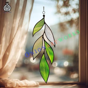 May include: A stained glass leaf decoration with a black outline. The leaves are various shades of green and white. The decoration hangs from a small metal ring. The background is a blurred view of a window and curtains.