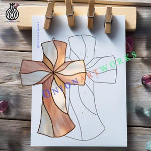 May include: A stained glass cross with a copper frame, featuring a design of swirling colors including white, peach, and tan. The cross is displayed on a white paper with a cross outline and held by wooden clips. The text "@OnionArtWorks" is visible.