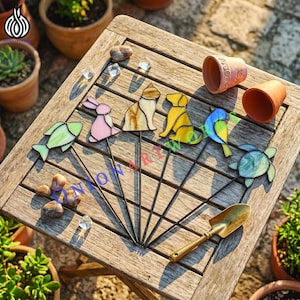 May include: A collection of colorful stained glass garden stakes in the shapes of a fish, rabbit, cat, dog, bird, and turtle. The stakes are arranged on a wooden table with a small trowel and some decorative stones.
