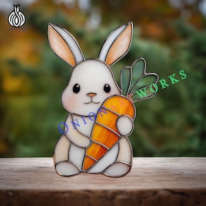 Może przedstawiać: Witrażowy królik trzymający dużą pomarańczową marchewkę z zielonymi liśćmi. Królik jest biały z pomarańczowymi uszami i policzkami. Obrazek zawiera tekst "ONION ART WORKS".