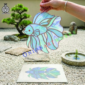 Puede incluir: Un pez betta de vidriera en tonos azules y verdes, con contornos de color cobre. El pez está suspendido por una cadena dorada. La imagen incluye el texto "ONION ART WORKS".