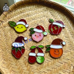 Op de afbeelding: Zes glas-in-lood fruitornamenten in de vorm van een citroen, perzik, appel, aardbei, kersenpaar en sinaasappel. Elk stuk is voorzien van een feestelijke rood-witte kerstmuts, een groen blaadje en een eenvoudig lachend gezichtje met oogjes en mond.