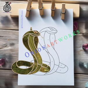 May include: A stained glass cobra in green and gold, with an orange eye and a forked tongue. The cobra is on white paper with a line drawing of a cobra. The paper is held by wooden clips.