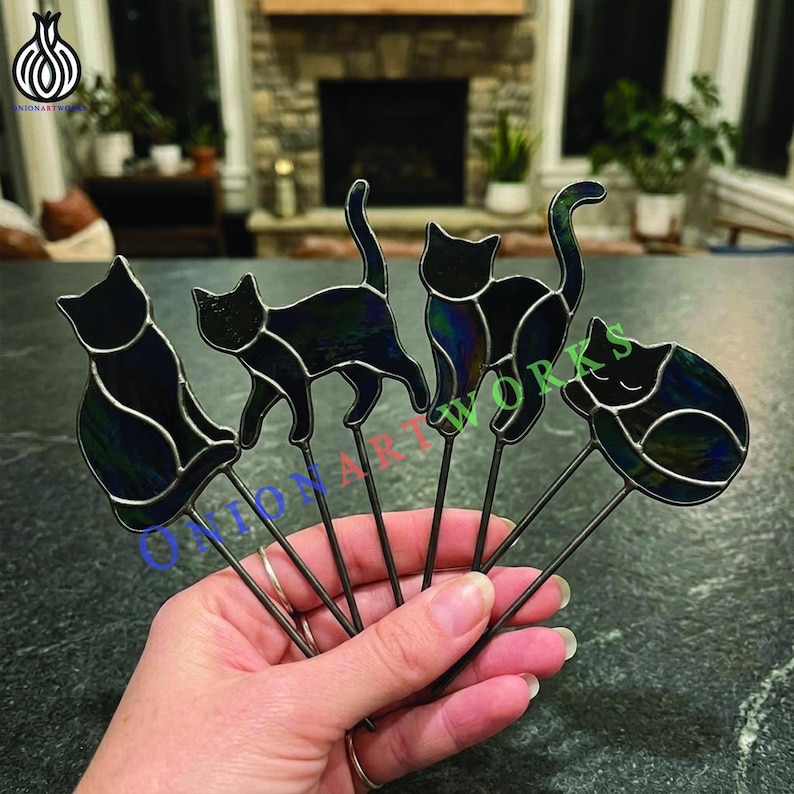 May include: Four stained glass cat ornaments in various poses, each featuring dark, iridescent glass with metallic solder lines. The cats are attached to thin metal rods. The text ONIONARTWORKS is overlaid in blue, red, and green across the center.