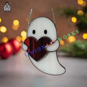 May include: A white stained glass ghost holding a red heart. The ghost has black eyes and is suspended by a silver chain. The text "ONION ART WORKS" is visible on the heart. The background features blurred lights and red ornaments.