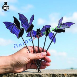 May include: A set of four stained glass bat ornaments featuring purple wings with black glass bodies and edges. Each bat is attached to a thin metal rod, creating a cohesive decorative display with translucent purple glass textures.