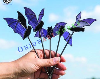 May include: A set of four stained glass bat ornaments featuring purple wings with black glass bodies and edges. Each bat is attached to a thin metal rod, creating a cohesive decorative display with translucent purple glass textures.