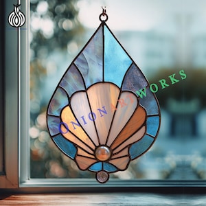 以下が含まれることがあります： 涙の形をしたステンドグラスの貝殻サンキャッチャー。デザインは、クリーム色、タン色、白のセグメントを持つ中央の貝殻と、青とティールのガラスで囲まれています。真珠のビーズと小さな透明なガラスビーズが一番下にあります。