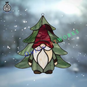 May include: A stained glass suncatcher shaped like a Christmas tree with a gnome in the center. The gnome has a dark red hat, a white beard, and brown boots. The tree is made of green glass panels with black lead lines and a metal hanging loop.