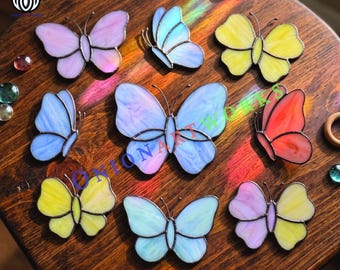 May include: A collection of nine stained glass butterflies in various colors including pink, yellow, blue, and red, each featuring dark metal soldering lines. The butterflies are arranged on a dark wood surface with the text ONIONARTWORKS visible.