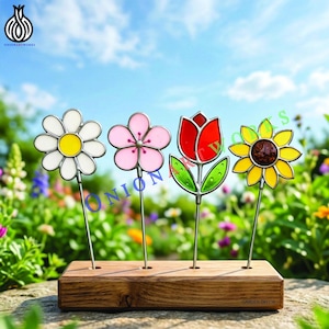 Op de afbeelding: Vier kleurrijke glazen bloemtuinstokken. De bloemen zijn een madeliefje, een roze bloem, een rode tulp en een gele zonnebloem. De stokken staan in een houten voet. De tekst "GARDEN DECOR" is zichtbaar.