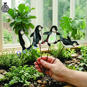May include: Four stained glass penguin ornaments on thin metal stakes. Each penguin features a black and white design with iridescent glass panels. The penguins are depicted in various poses, including standing and swimming, connected to metal rods.