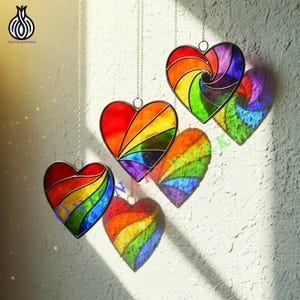 Könnte beinhalten: Bunte Glas-Herz-Suncatcher in Regenbogenfarben, an silbernen Ketten aufgehängt. Jedes Herz hat ein einzigartiges Design mit leuchtenden Rot-, Orange-, Gelb-, Grün-, Blau- und Lilatönen. Die Suncatcher werfen farbige Schatten.