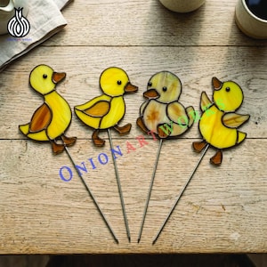 以下が含まれることがあります： 金属製の杭に取り付けられた4つのステンドグラス製のアヒルのオーナメント。3羽は明るい黄色に茶色のアクセントがあり、1羽は淡いまだら模様の黄色です。それぞれのアヒルには小さな黒い目があり、全体に暗い金属のハンダ付けラインが施されています。
