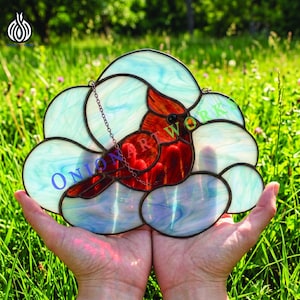 Puede incluir: Vidriera con un pájaro cardenal rojo. El ave se posa sobre un fondo en forma de nube con tonos azules y blancos. La pieza tiene un marco de metal oscuro y una cadena para colgar. Mide aproximadamente 20 cm.