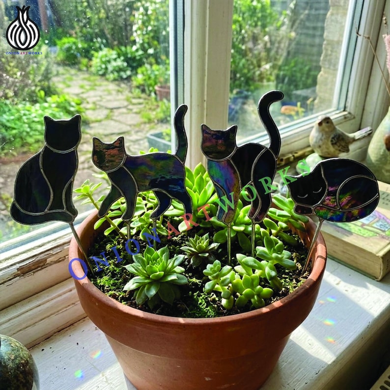 May include: Four stained glass cat silhouettes on metal stakes inserted into a terracotta pot filled with green succulents. The cats feature iridescent, dark, and reflective glass panels with soldered metal seams, displaying various poses.