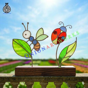 Op de afbeelding: Een gebrandschilderde glazen sculptuur met een bij en een lieveheersbeestje, elk met kleurrijke details. De bij heeft een witte kop, blauwe ogen en een oranje lichaam. Het lieveheersbeestje is rood met zwarte stippen. Beide zijn gemonteerd op een houten voet.