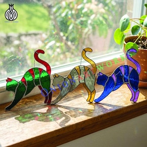 Op de afbeelding: Drie glas-in-lood kattenvangers in groen, goud en blauw met gebogen ruggen en gebogen staarten. Elke kat heeft een ander kleurenschema en hangt aan een ketting. De katten worden tentoongesteld op een houten vensterbank.