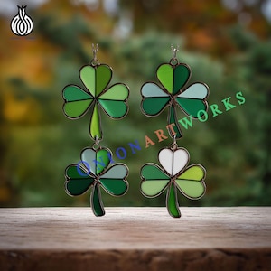 May include: Four shamrock earrings with a bronze-colored frame. Each shamrock is divided into three sections of varying shades of green and white. The earrings are hanging against a blurred green background.
