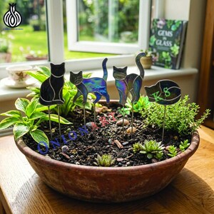 May include: Four stained glass cat garden stakes are inserted into a large, round, terracotta-colored planter filled with soil and various green plants. The cats feature iridescent, blue, purple, and black glass panels with thin metal solder lines.